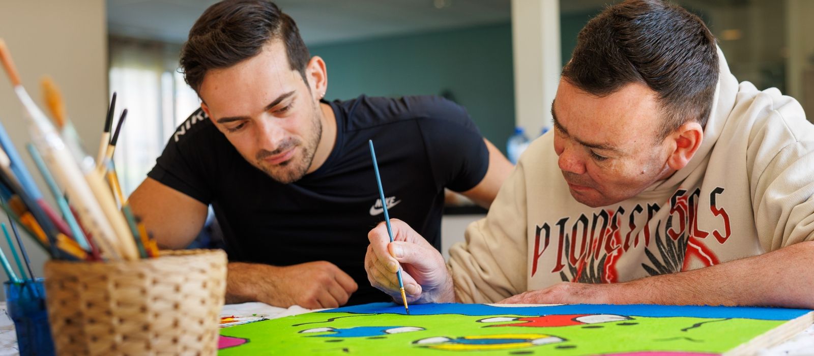 Een begeleider en een cliënt zijn samen aan het verven, waarbij zij aandachtig naar een groot beschilderd vel papier kijken met een penseel in de hand.