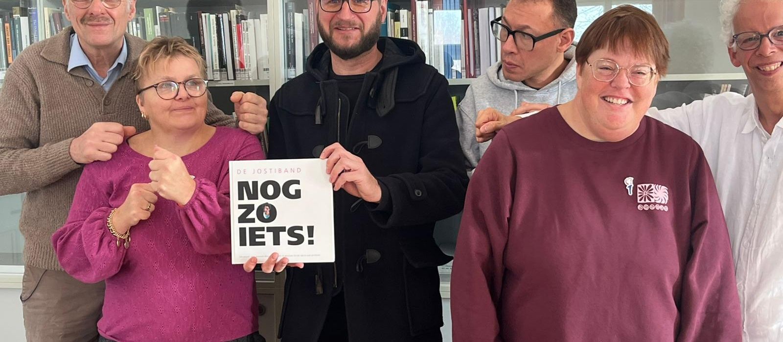 Vijf Jostibandleden en begeleiders staan in een rij naast elkaar voor een grote boekenkast. Ze houden een boek vast met de titel "De Jostiband: nog zoiets".