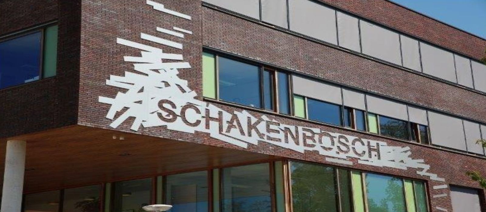 Een wit bord geplaatst boven de hoofdingang van het pand Schakenbosch met in donkerblauwe letters 'Schakenbosch.'