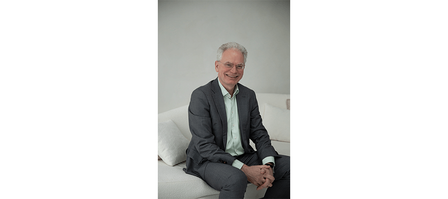 Meneer met bril zit in pak op een donkergekleurde bak en kijkt glimlachend in de camera