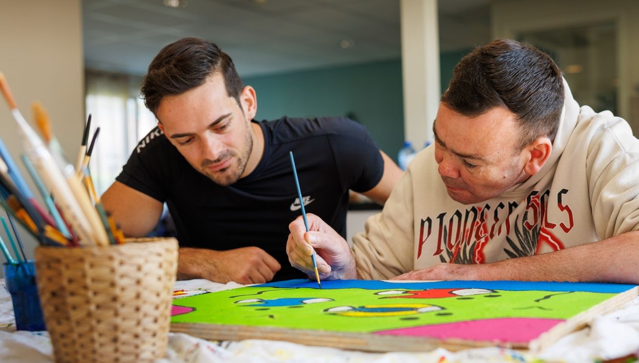Een begeleider en een cliënt zijn samen aan het verven, waarbij zij aandachtig naar een groot beschilderd vel papier kijken met een penseel in de hand.